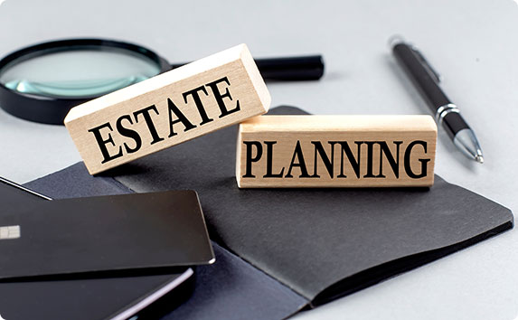 Wooden blocks spell out "estate planning" on a notebook. Magnifying glass and pen nearby.